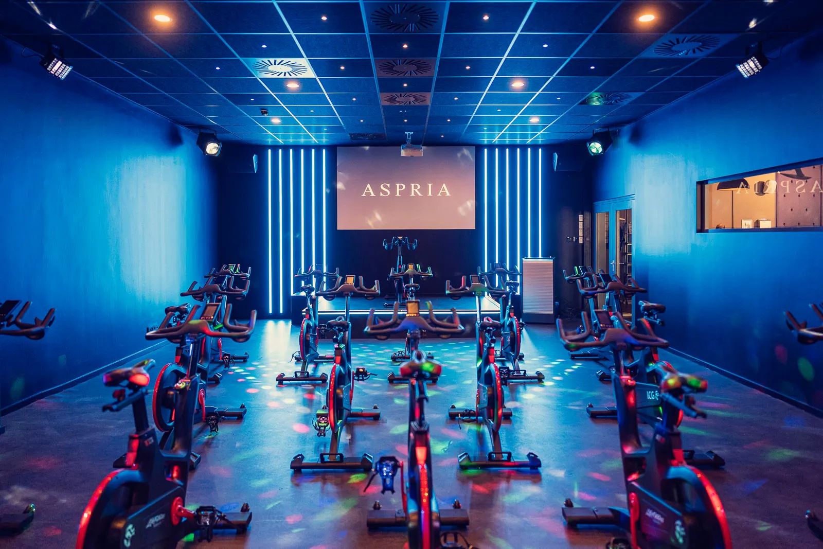 Leerer Indoor-Cycling-Raum mit Spinningrädern in Reihen, blauen Lichtleisten und bunten Lichtpunkten auf dem Boden.
