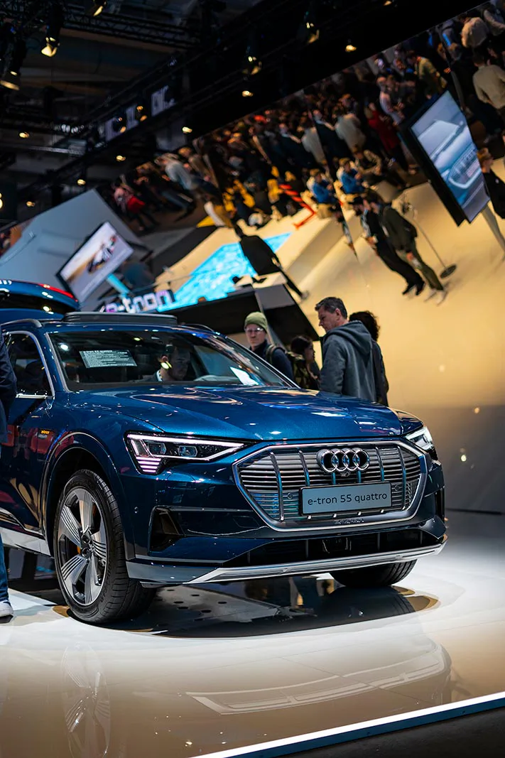 Blauer Audi e-tron auf beleuchtetem Messestand, dahinter Besucher und eine große Spiegelwand.