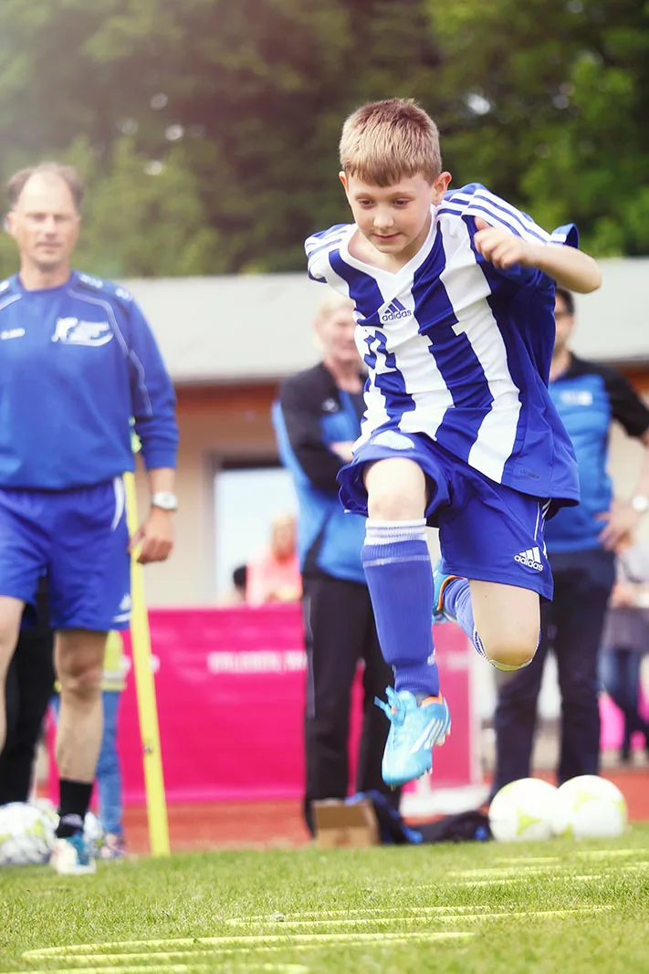 Junge im blau-weißen Fußballtrikot springt auf dem Rasen über eine Trainingsleiter, Erwachsene schauen zu.
