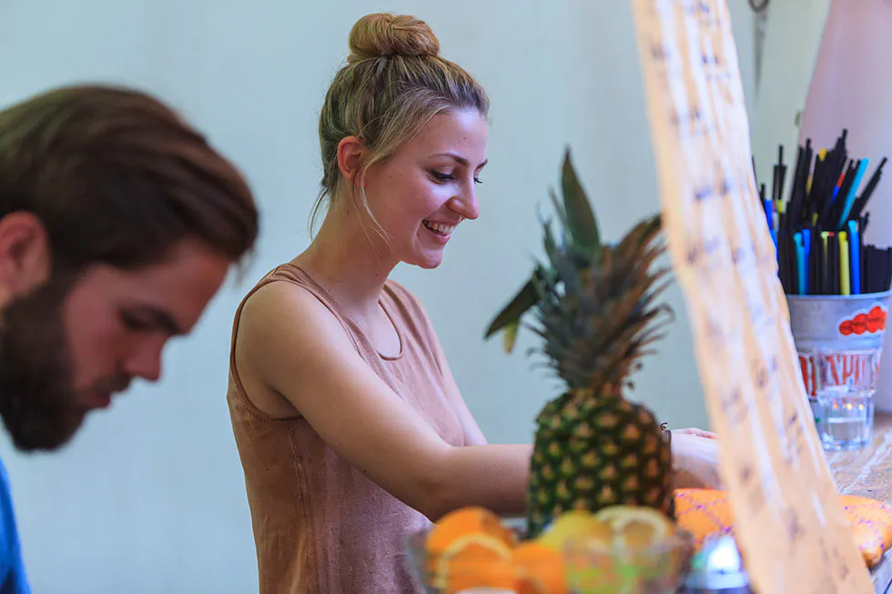 Lächelnde Frau hinter einem Getränkestand mit Ananas, Zitrusfrüchten und Strohhalmen; links ein bärtiger Mann.