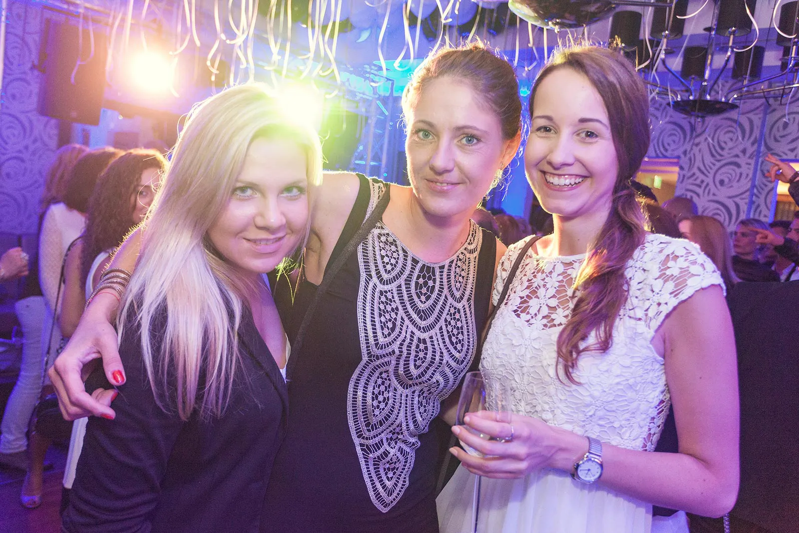 Drei Frauen posieren bei buntem Partylicht unter Luftschlangen, eine hält ein Glas in der Hand.