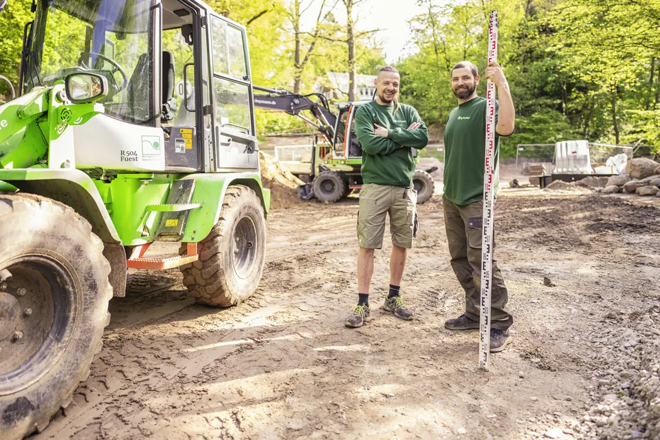 Zwei Männer in grünen Oberteilen stehen neben einem Radlader auf einer sonnigen Baustelle, einer hält eine Messlatte.