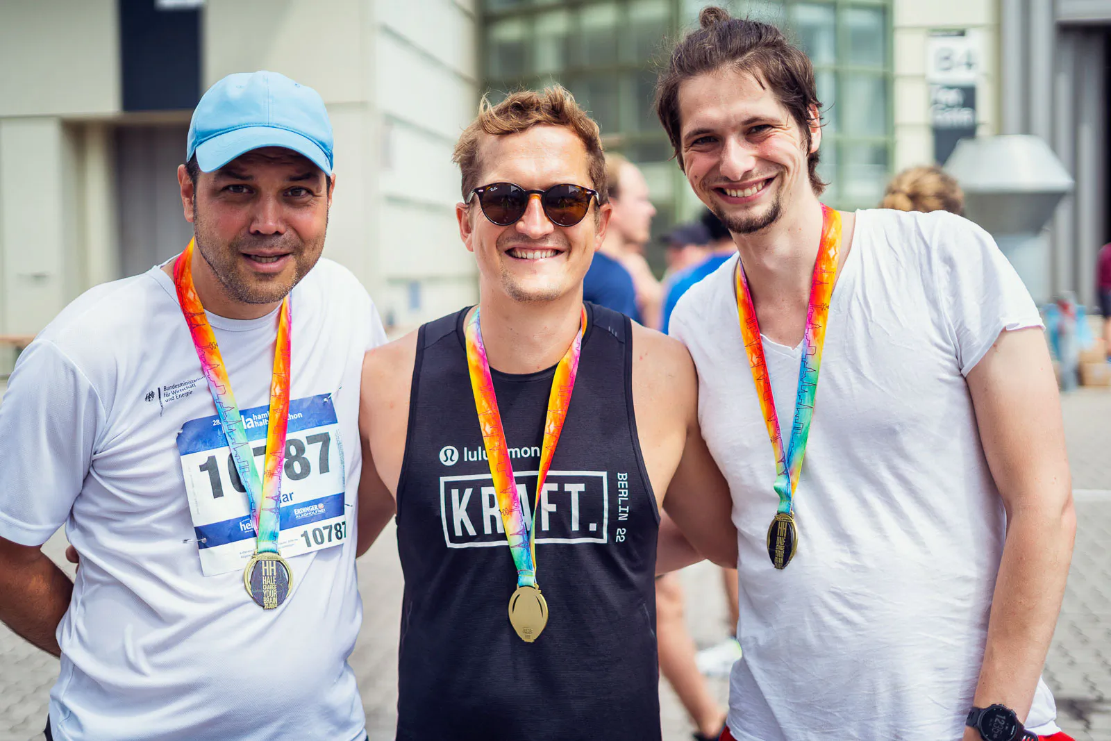Drei lächelnde Läufer mit Medaillen posieren beim Hella Halbmarathon vor einem Gebäude im Tageslicht.