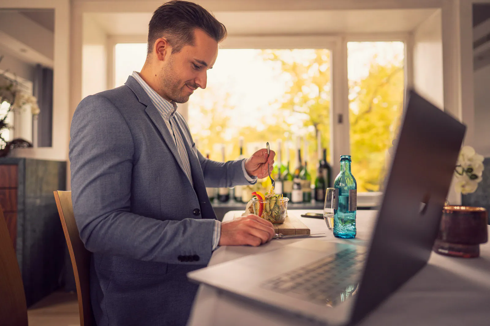 Mann im blauen Sakko isst am Tisch neben offenem Laptop und Wasserflasche, im warmen Tageslicht großer Fenster.
