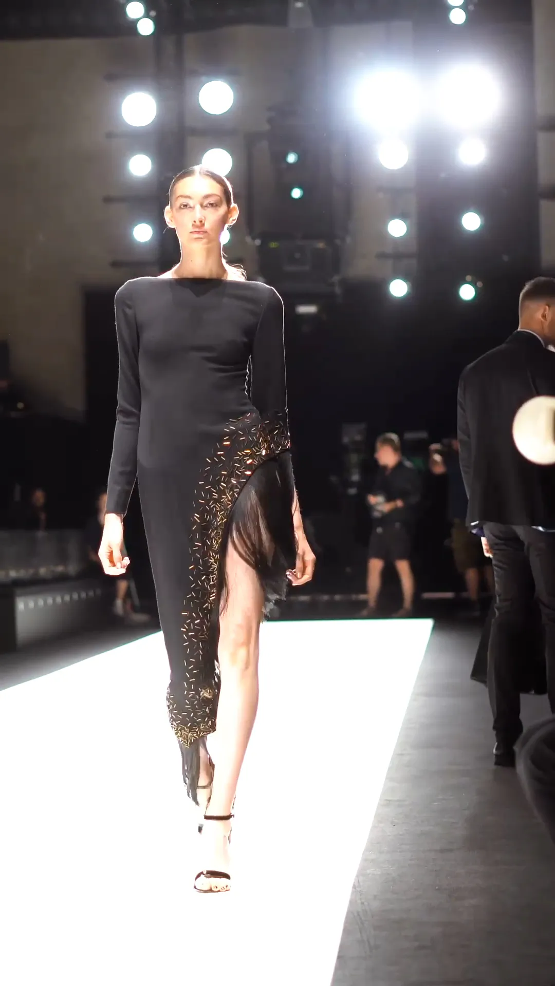 Model im schwarzen Kleid mit hohem Beinschlitz läuft unter hellen Scheinwerfern über den Laufsteg.
