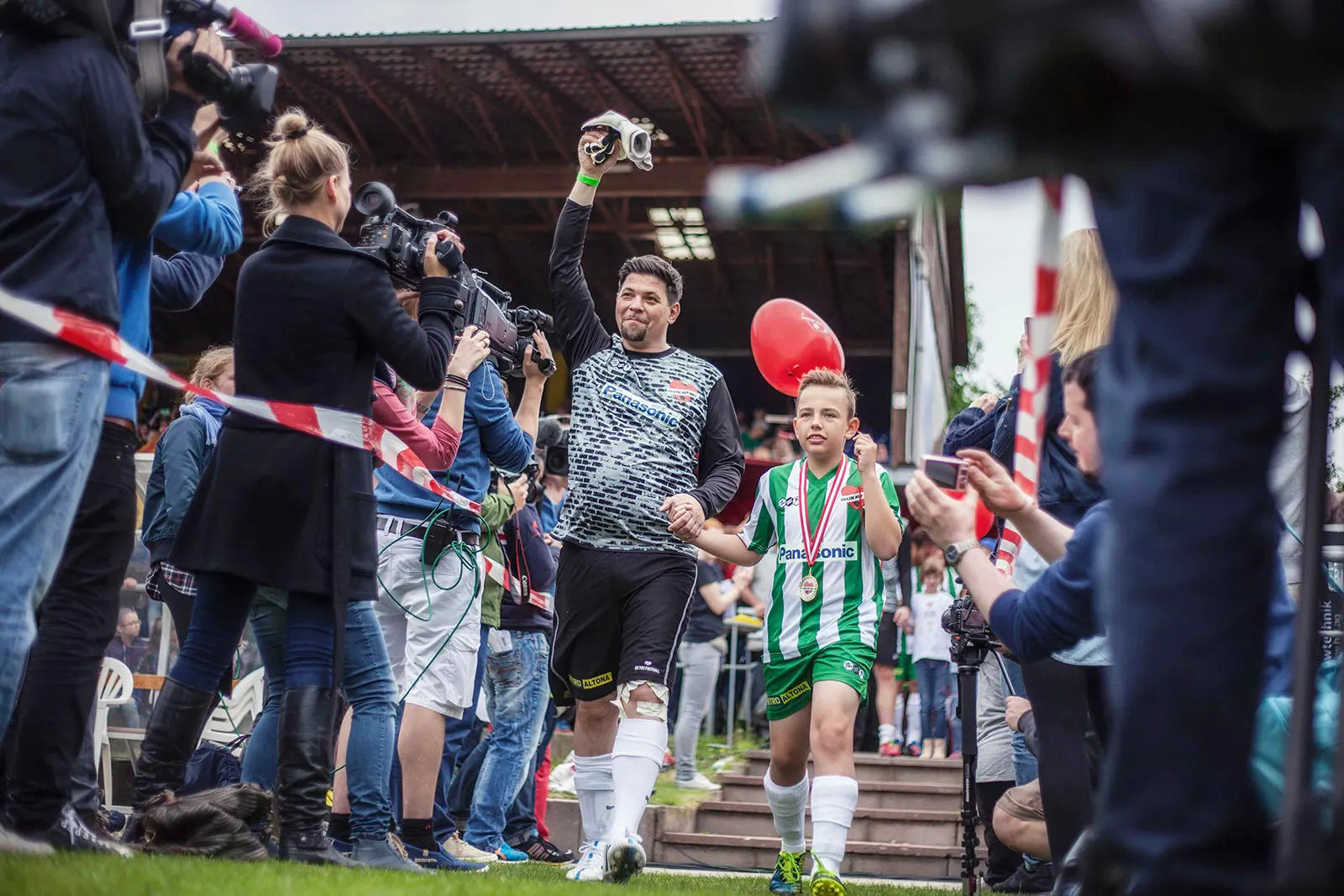 Fußballspieler und Kind im grün-weißen Trikot laufen zwischen Kamerateams und Zuschauern auf den Rasen.