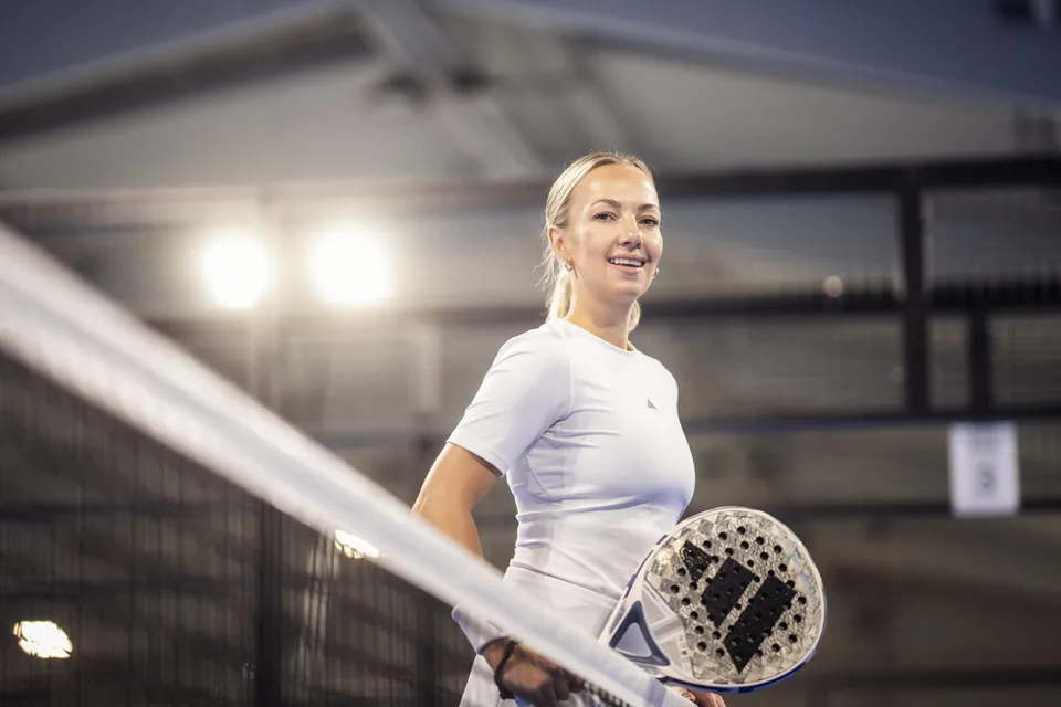 Lächelnde Frau im weißen Sportshirt mit Padelschläger am Netz eines Hallenplatzes im hellen Gegenlicht.