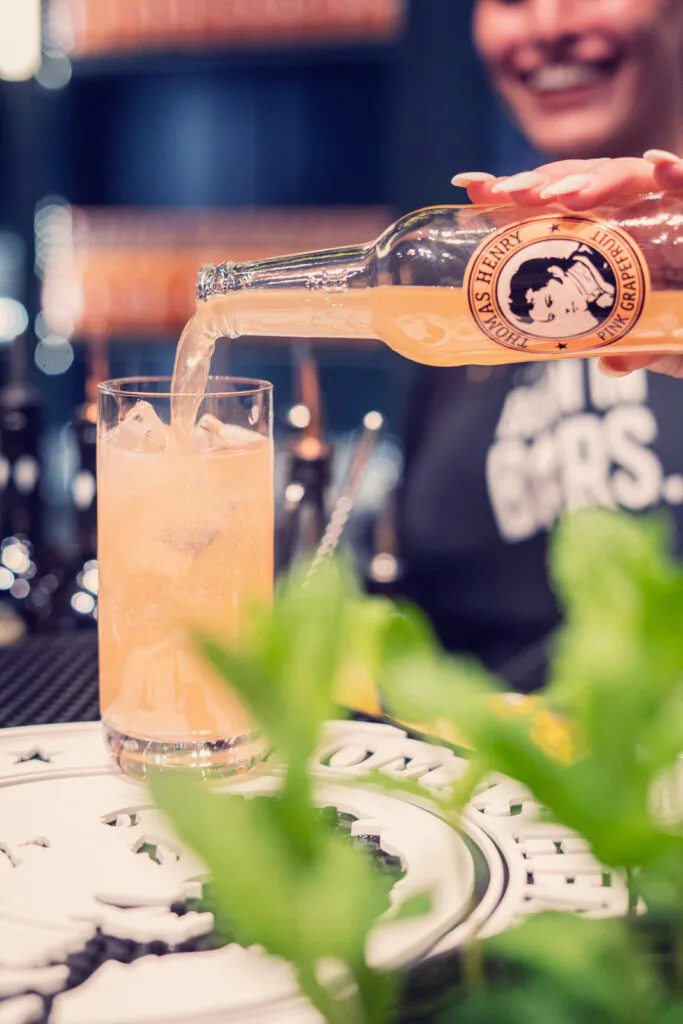 Lächelnde Person gießt Thomas Henry Pink Grapefruit aus einer Flasche in ein hohes Glas mit Eis an einer Bar.