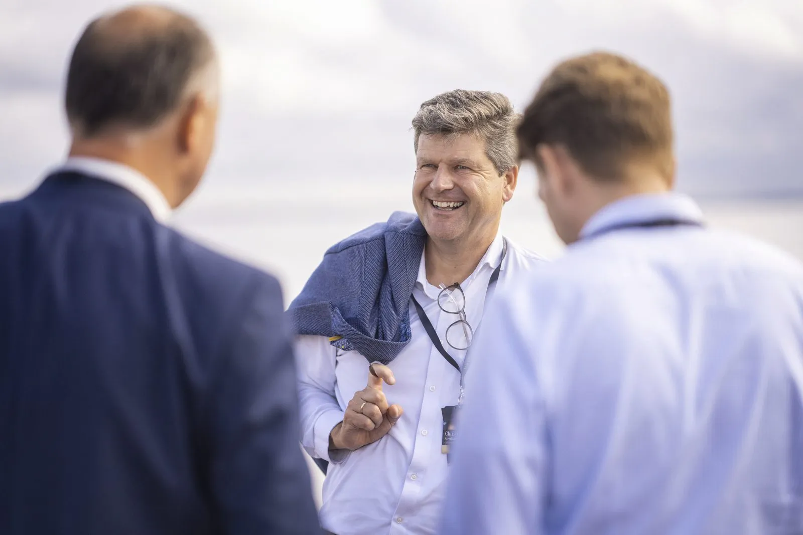 Drei Männer im Gespräch bei Tageslicht im Freien; der Mann in der Mitte lacht, mit einem Jackett über der Schulter.