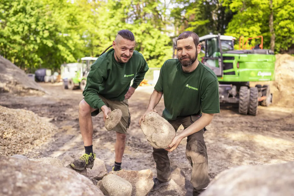 Zwei Boymann-Mitarbeiter halten große Steine auf einer sonnigen Baustelle, hinter ihnen steht ein grüner Bagger.