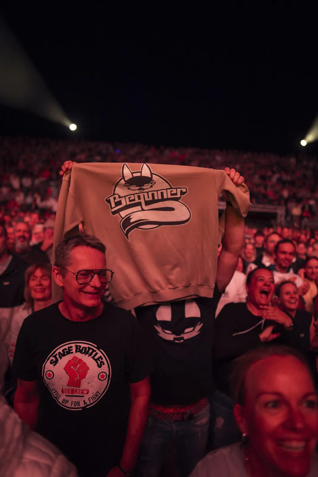 Ein Konzertbesucher hält einen beigen Pullover mit Beginner-Logo hoch, umgeben von Fans im roten Licht.