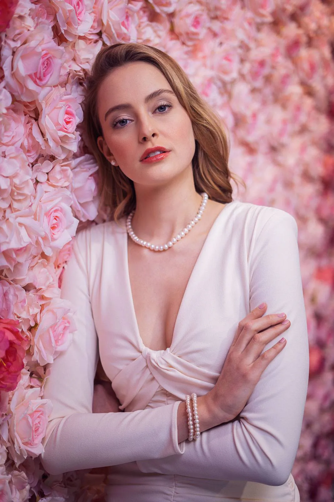 Frau im hellrosa Kleid mit Perlenkette und Perlenarmband vor einer rosa Blütenwand, weich beleuchtet.
