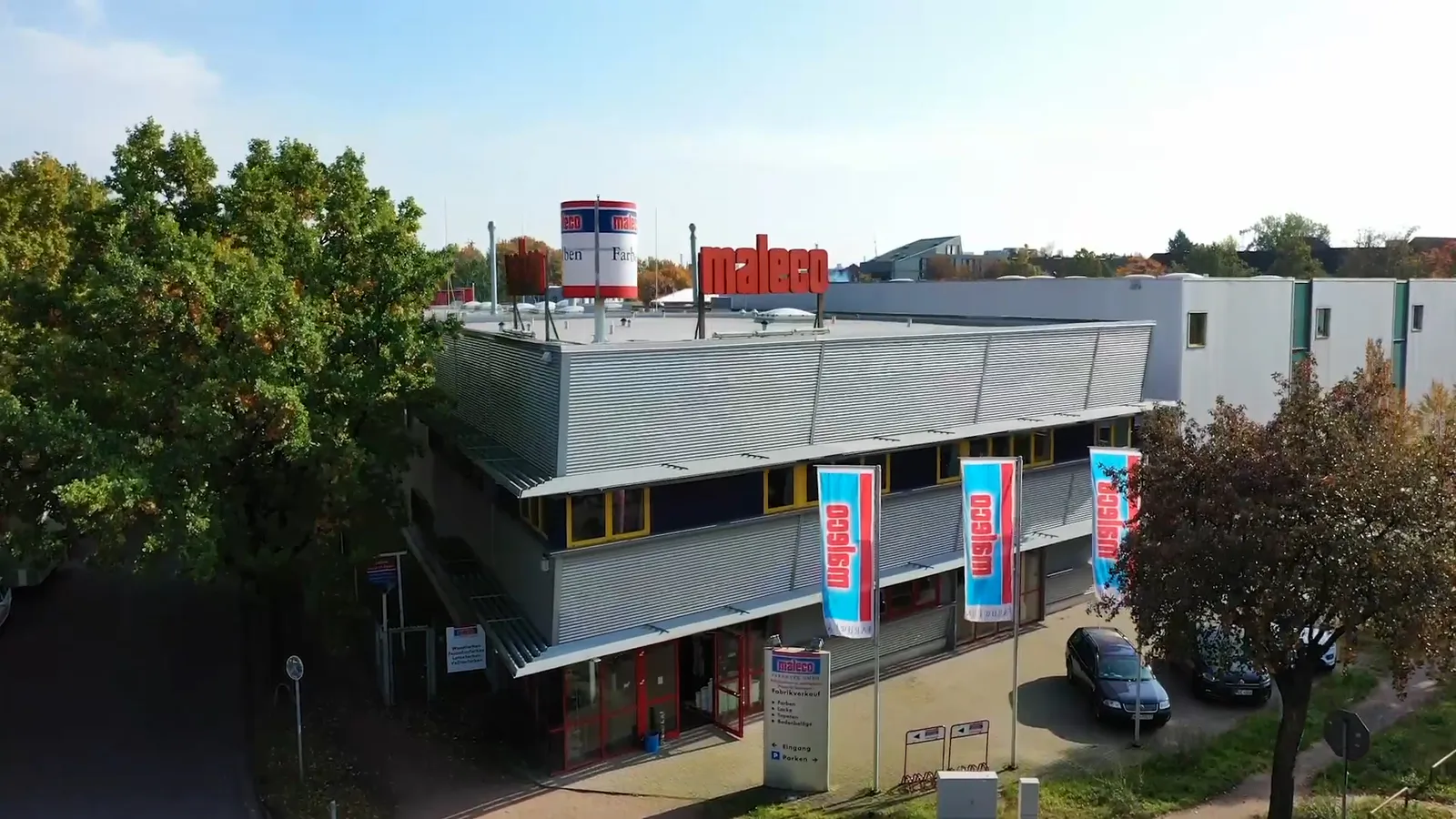 Maleco-Firmengebäude mit rotem Dachschriftzug, blauen Fahnen und parkenden Autos zwischen Bäumen bei Tageslicht.