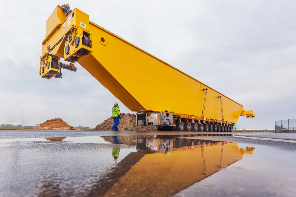 Gelber Kranträger auf Schwerlasttransporter, Arbeiter in Warnkleidung daneben; Spiegelung in Pfütze bei grauem Himmel.