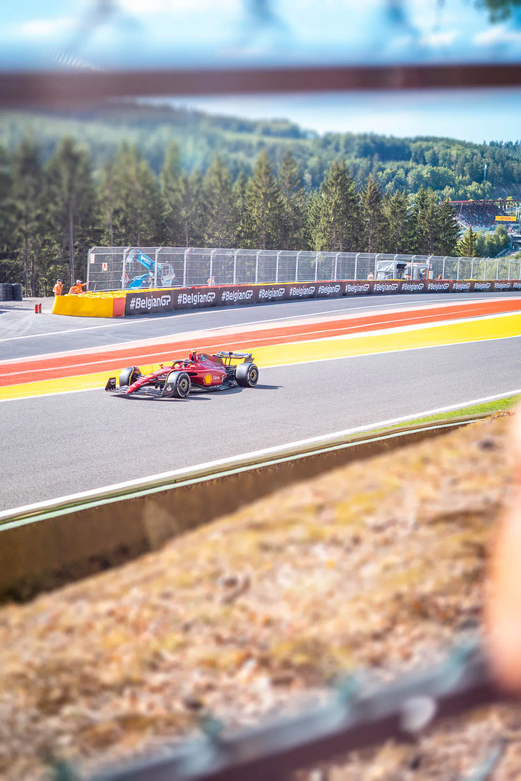 Roter Formel-1-Rennwagen auf sonniger Rennstrecke mit rot-gelben Randstreifen und bewaldeten Hügeln im Hintergrund.