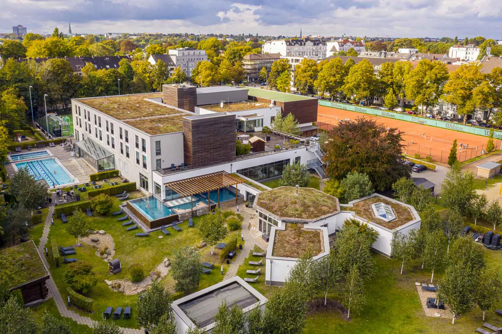 Drohnenansicht der Aspria-Anlage mit Außenpools, Tennisplätzen, begrünten Dächern und Liegewiese bei Tageslicht.