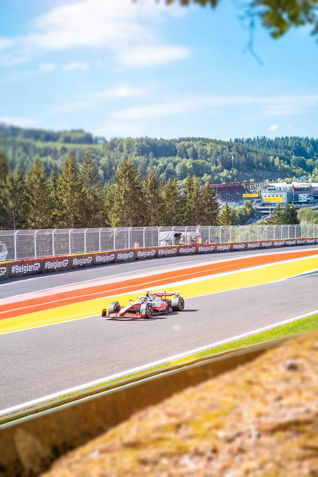 Haas-F1-Rennwagen auf sonniger Rennstrecke mit rot-gelben Randstreifen vor bewaldeten Hügeln.