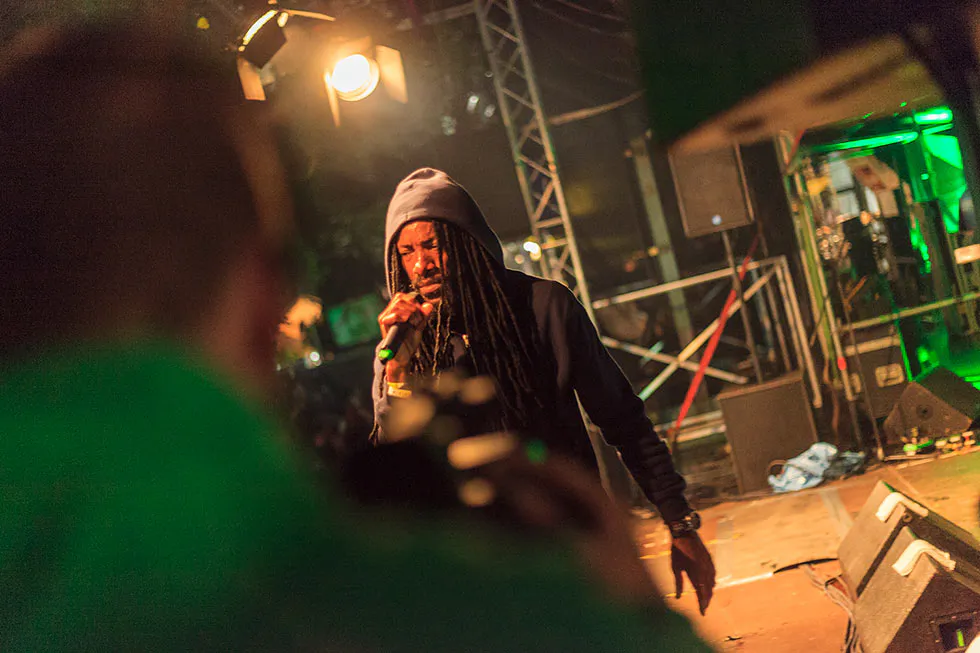 Sänger mit Dreadlocks und Kapuze singt ins Mikrofon auf einer gelb-grün beleuchteten Bühne, davor eine unscharfe Person.