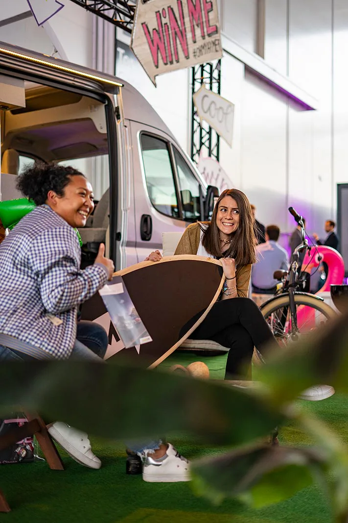 Zwei lachende Personen sitzen mit einem Surfbrett vor einem Camper in einer Messehalle, daneben ein Fahrrad.