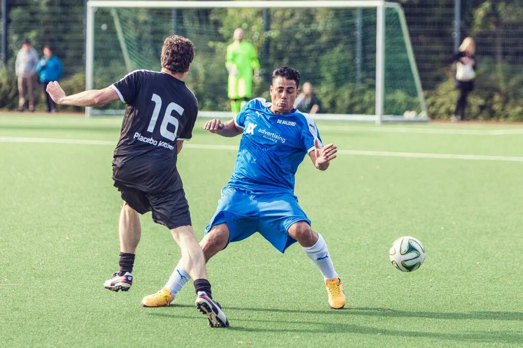 Zwei Fußballspieler in Blau und Schwarz kämpfen auf sonnigem Kunstrasen um den Ball, im Hintergrund ein Tor.