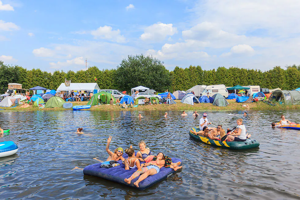 Festivalgäste schwimmen und treiben auf Luftmatratzen und Schlauchbooten im Wasser, dahinter Zelte am Ufer.