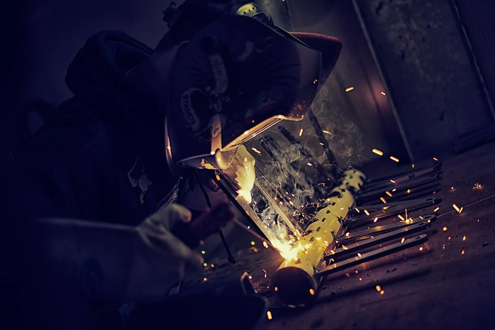 Schweißer mit Schutzhelm bearbeitet ein Metallrohr in dunkler Werkstatt, Funken und helles Schweißlicht leuchten auf.