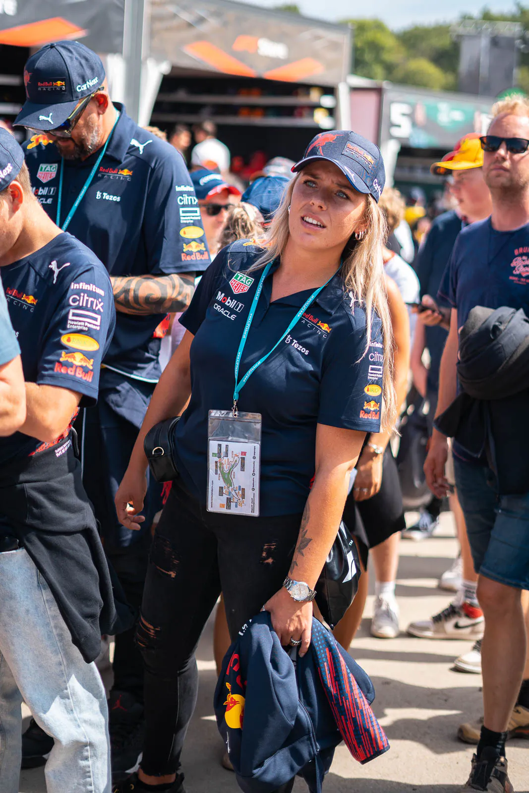 Frau in RedBull-Teamkleidung mit Kappe und Akkreditierung in einer Menschengruppe im sonnigen Fahrerlager.