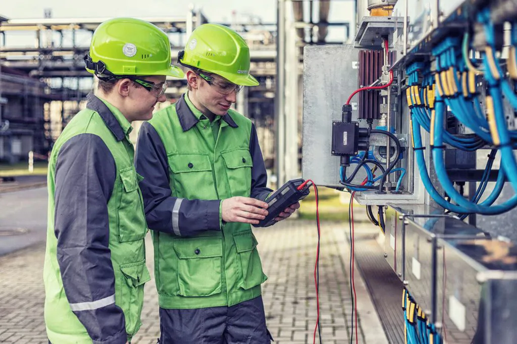 Zwei Arbeiter mit grünen Helmen prüfen mit einem Messgerät eine Elektroanlage mit blauen Kabeln in der Raffinerie Heide.