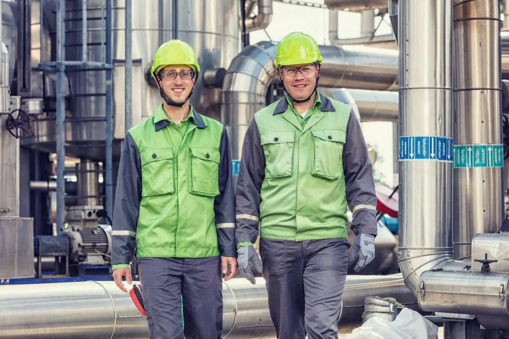 Zwei Mitarbeiter in grüner Schutzkleidung mit Helmen und Schutzbrillen zwischen Metallrohren der Raffinerie Heide.