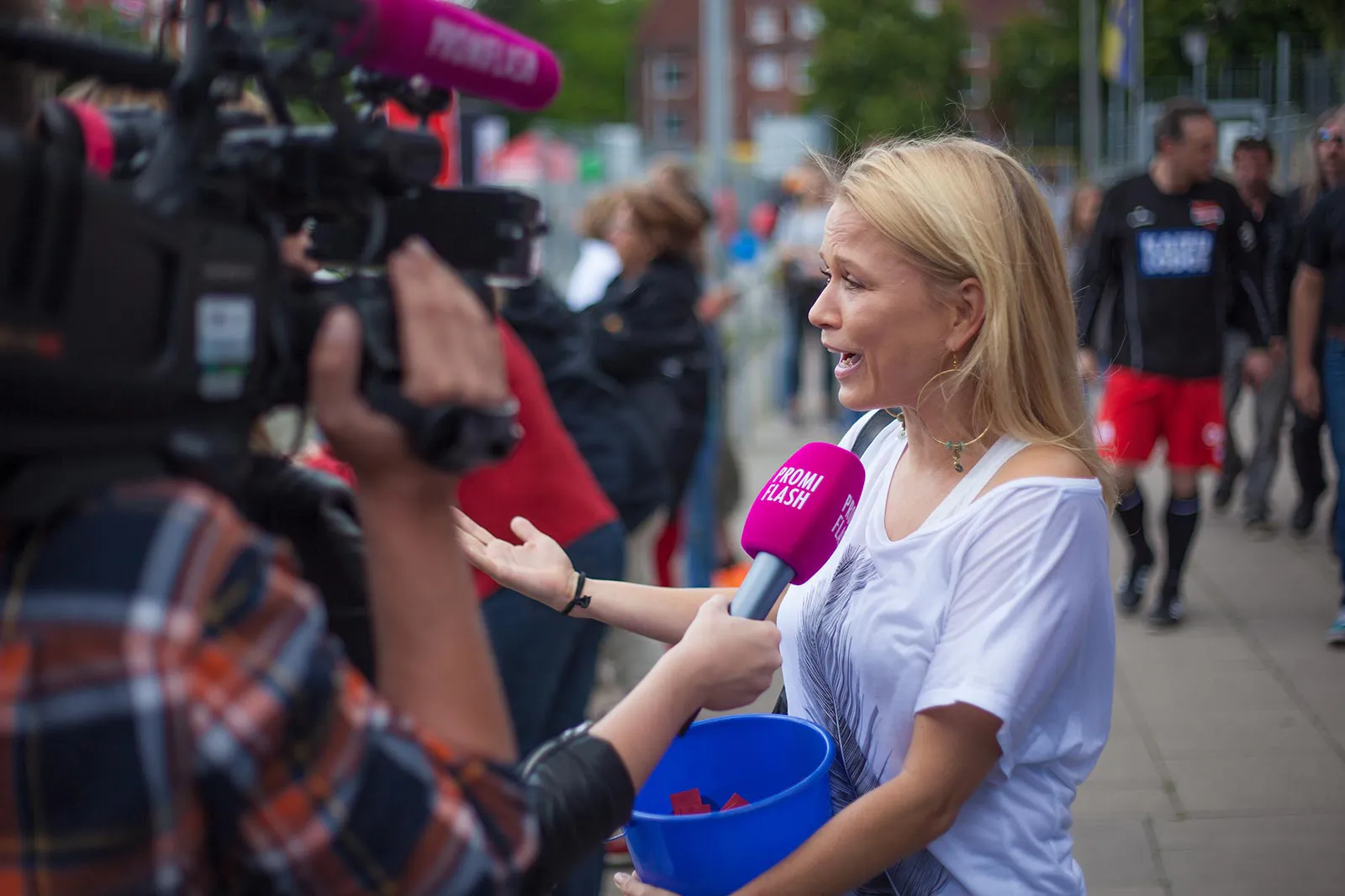 Blonde Frau mit blauem Eimer gibt draußen ein Interview vor einer Kamera und pinkem Mikrofon.