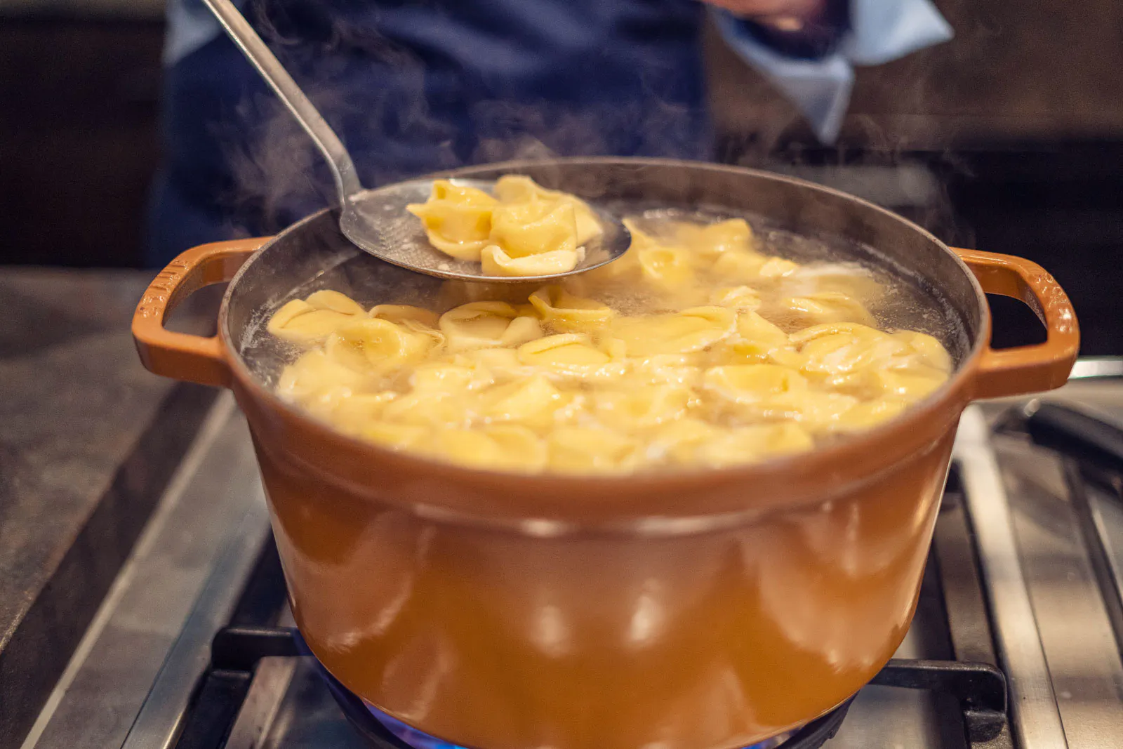 Gefüllte Pasta kocht in einem orangefarbenen Topf auf dem Gasherd, eine Schaumkelle hebt Nudeln aus dem Dampf.