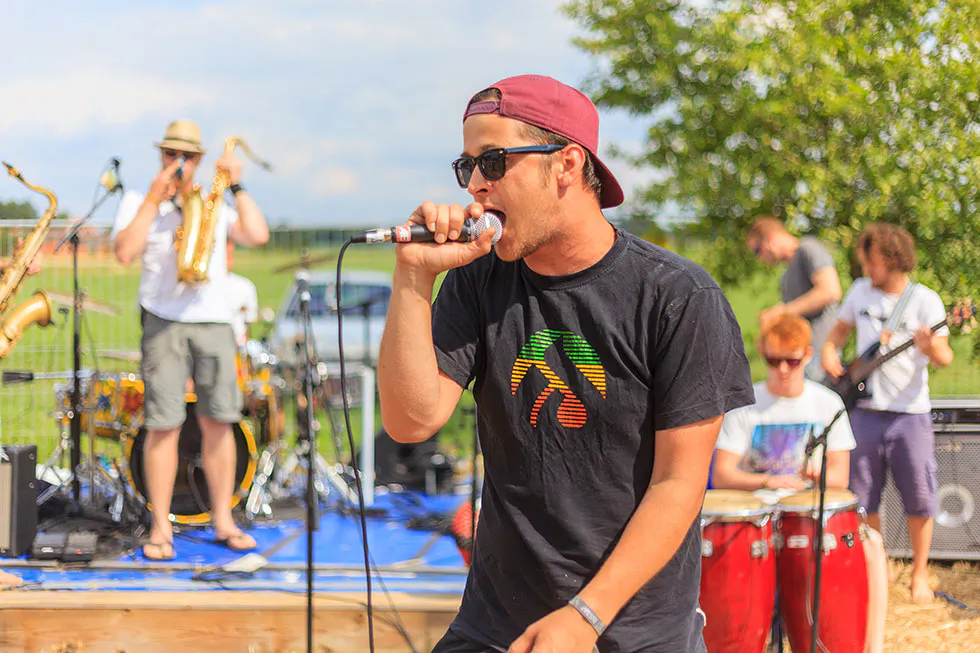 Sänger mit roter Kappe und Sonnenbrille singt ins Mikrofon, dahinter eine Band auf sonniger Freiluftbühne.