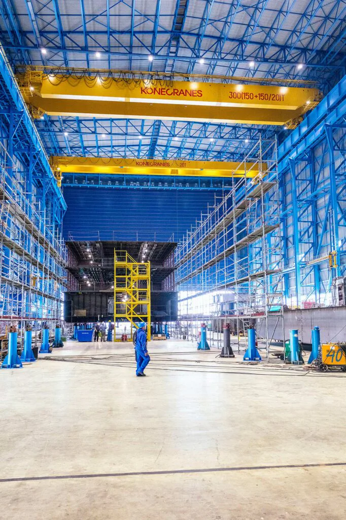 Arbeiter in Blau unter gelben Konecranes-Brückenkranen in einer hohen, hell beleuchteten Industriehalle.
