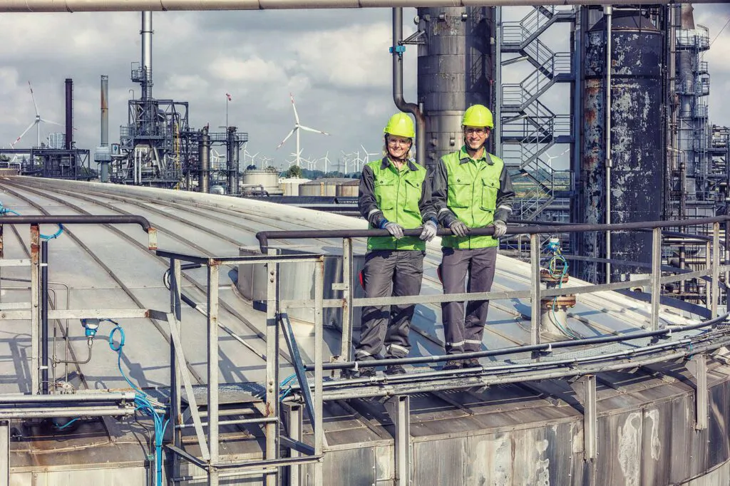 Zwei Mitarbeiter mit hellgrünen Helmen und grüner Schutzkleidung auf einer Plattform zwischen Anlagen der Raffinerie Heide.