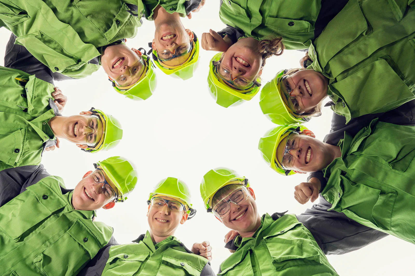 Personen in grüner Arbeitskleidung, Helmen und Schutzbrillen blicken im Kreis von oben in die Kamera.