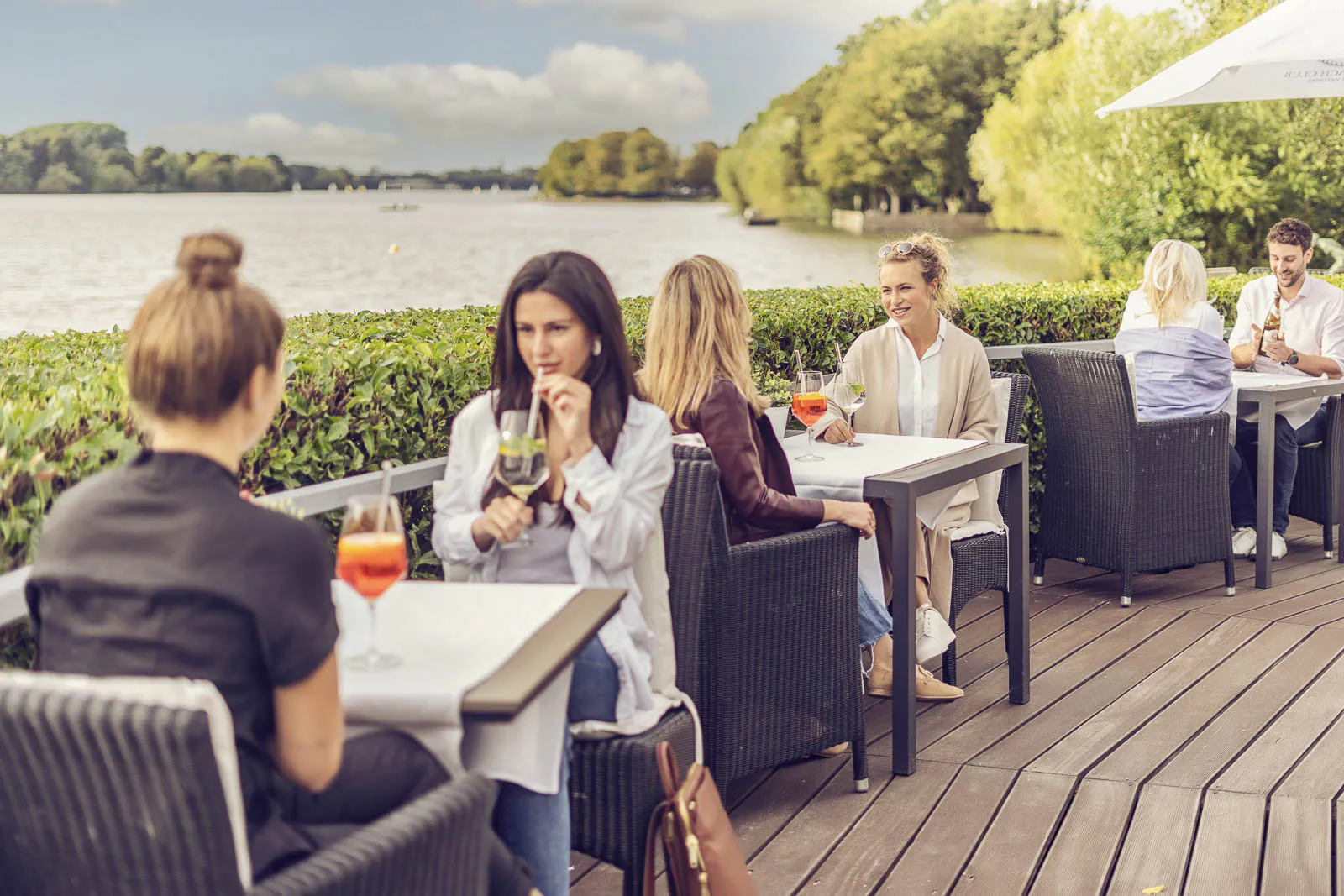 Gäste sitzen mit bunten Getränken an Tischen auf der sonnigen Holzterrasse des Insel Beach Club Hannover am See.
