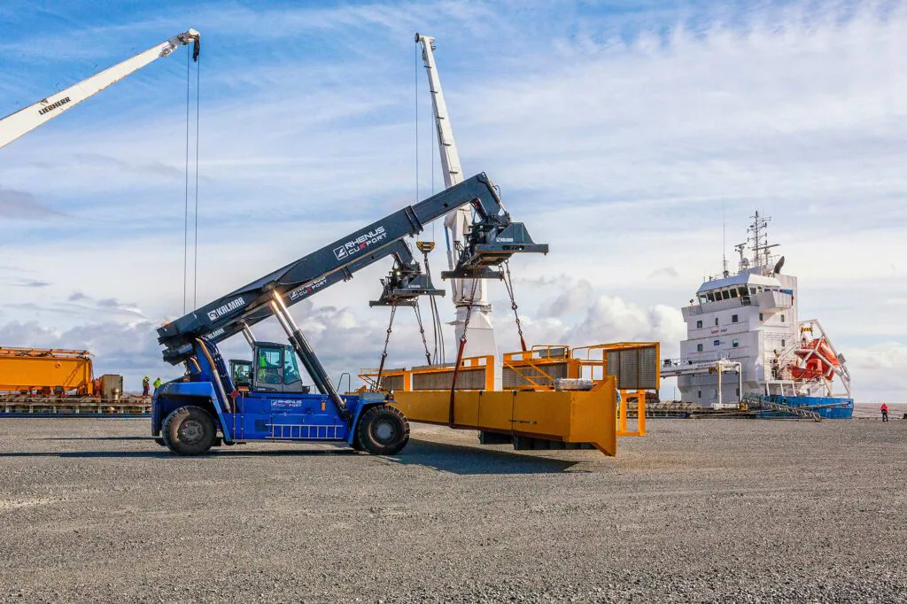 Blauer Reachstacker hebt ein gelbes Stahlbauteil am Hafenkai bei Tageslicht, dahinter Kräne und ein Schiff.