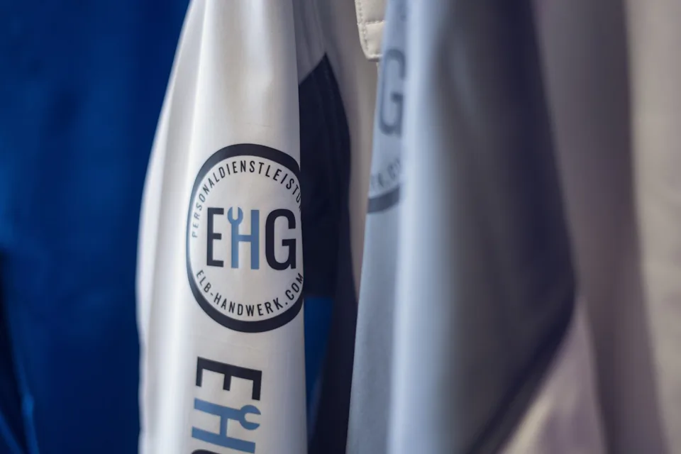 Nahaufnahme hängender Arbeitsjacken in Weiß, Grau und Blau mit EHG-Logo und Elb-Handwerk-Schriftzug.