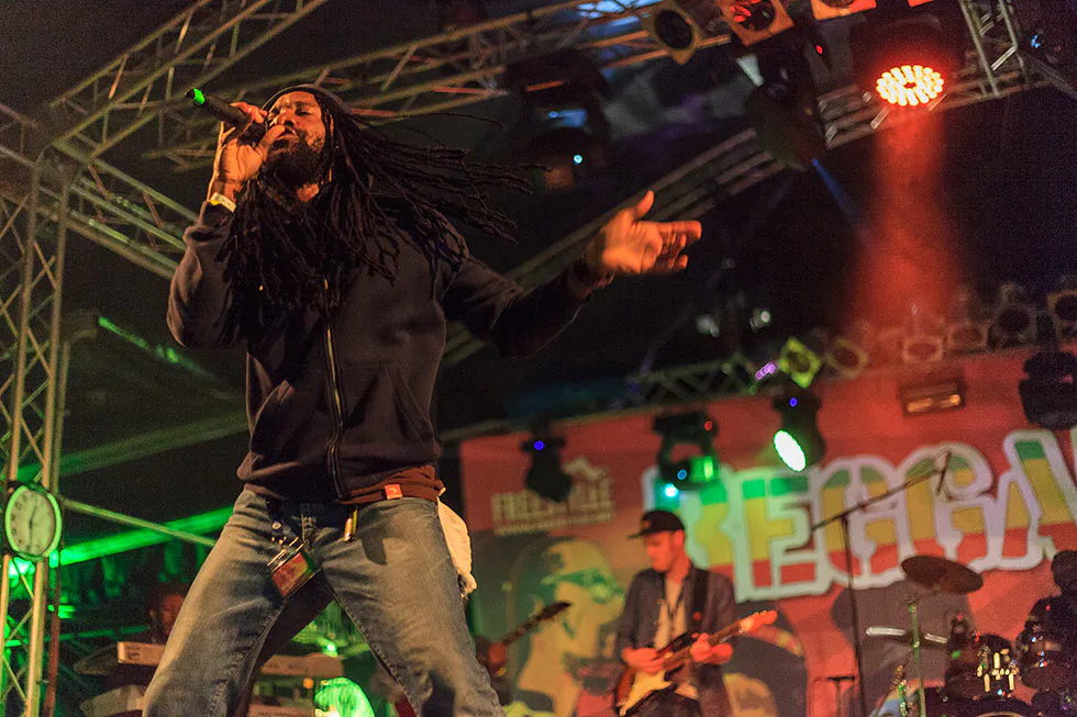 Sänger mit langen Dreadlocks singt ins Mikrofon, dahinter eine Band auf rot und grün beleuchteter Festivalbühne.