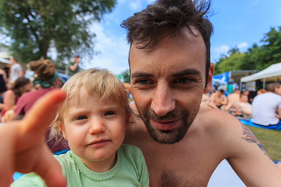 Kind greift zur Kamera neben einem lächelnden, oberkörperfreien Mann auf einer belebten Festivalwiese im Sonnenschein.
