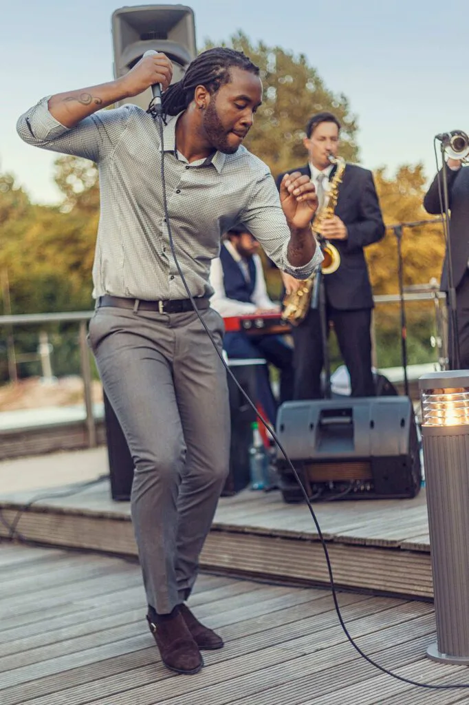 Sänger mit Mikrofon tanzt auf einer Holzterrasse, dahinter spielen Saxofonist und Keyboarder im Tageslicht.