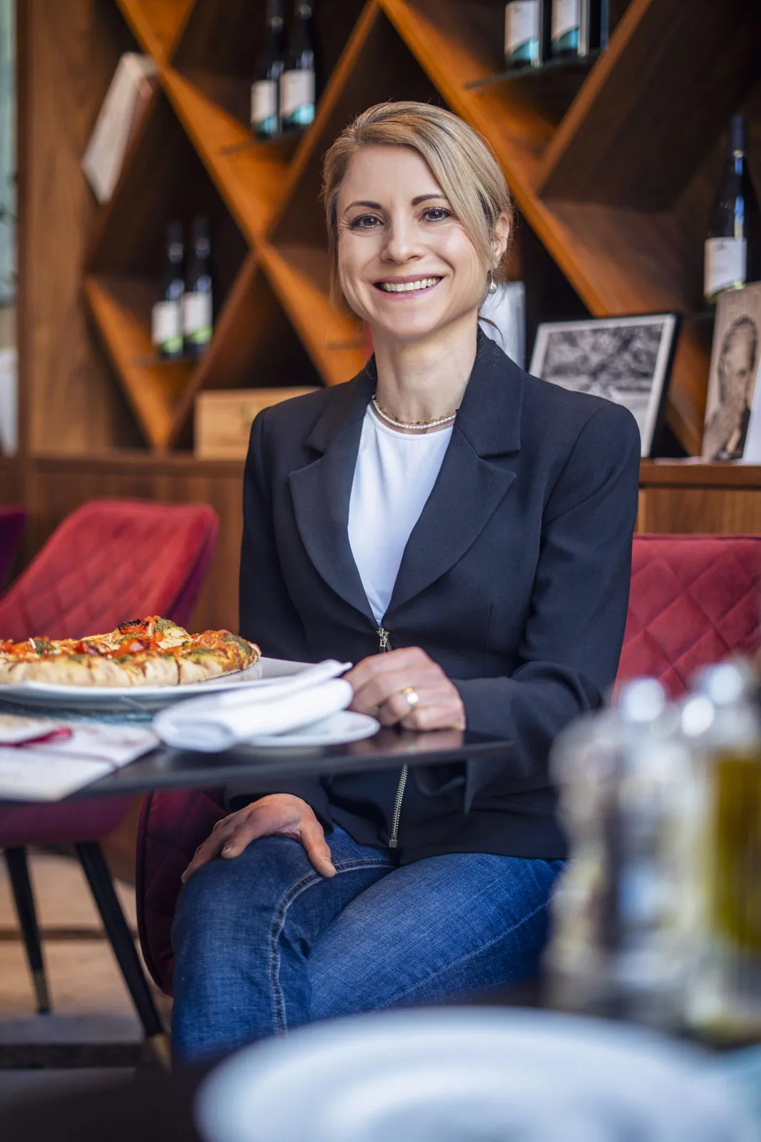 Lächelnde Frau im dunklen Blazer sitzt im Restaurant an einem Tisch mit Pizza, dahinter Weinflaschen im Holzregal.