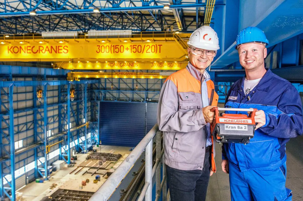 Zwei Arbeiter mit Schutzhelmen halten eine Kransteuerung vor gelben Konecranes-Brückenkranen in einer Industriehalle.