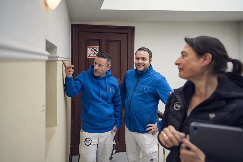 Zwei Handwerker in Blau und eine Frau mit Tablet im hellen Treppenhaus; ein Mann zeigt auf die Wand.