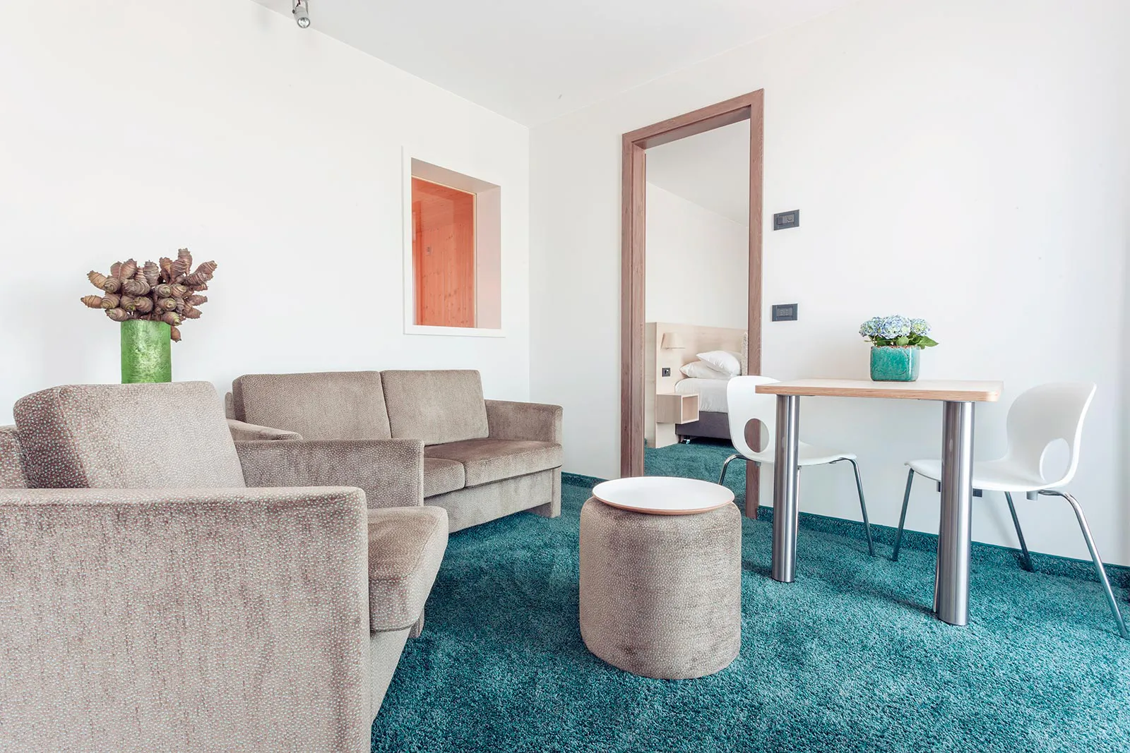 Heller Hotelwohnraum mit beigen Polstermöbeln, türkisfarbenem Teppich, Tisch und Blick ins Schlafzimmer.