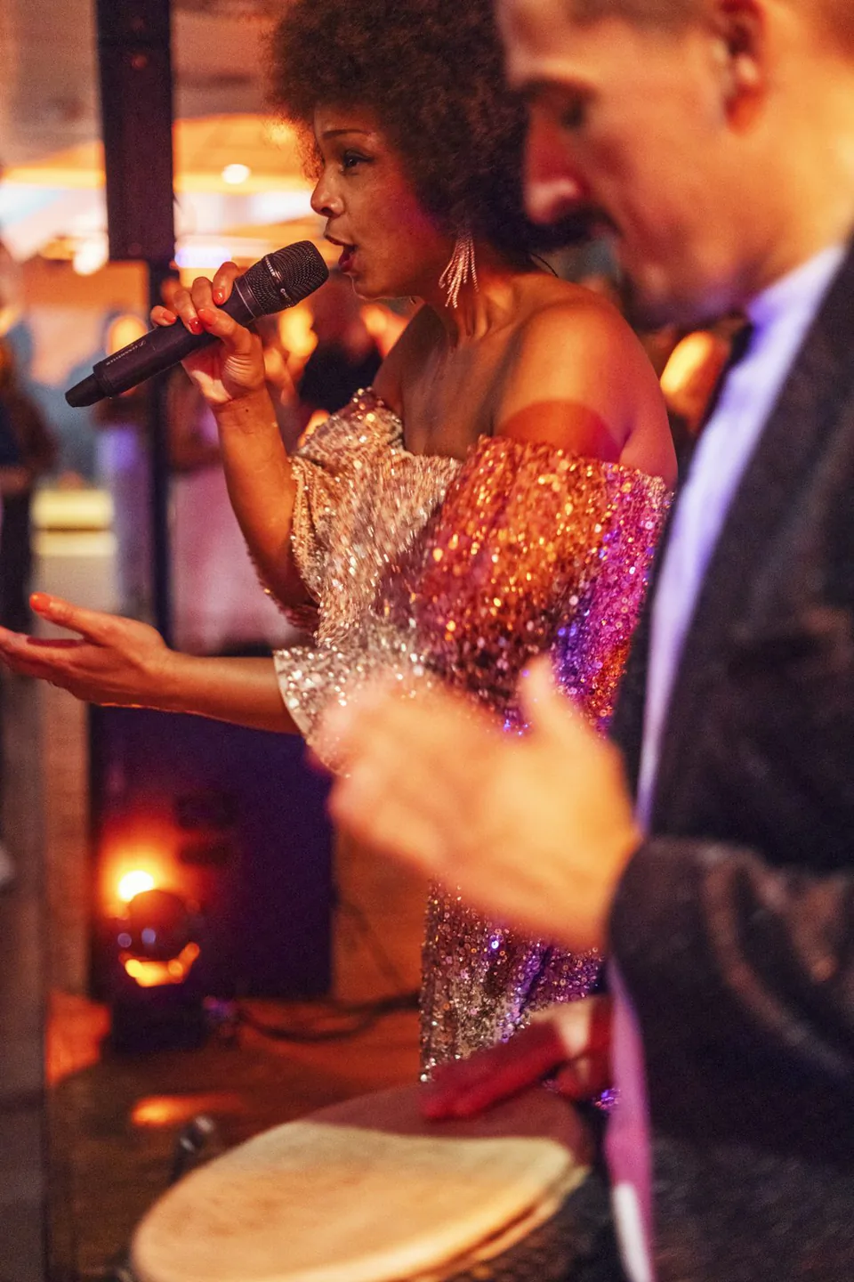 Sängerin im glitzernden Kleid mit Mikrofon neben einem Trommler bei warmem Licht auf dem Aspria-Event.