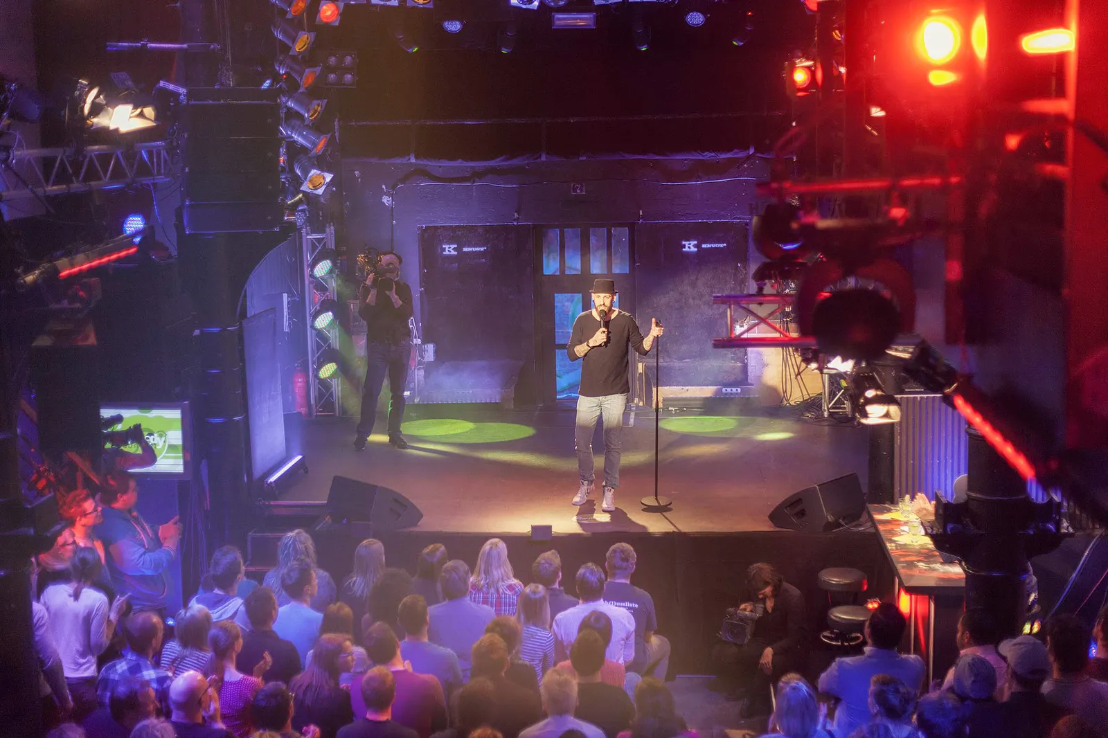 Dennis Grundt mit Mikrofon auf der bunt beleuchteten Bühne beim NDR Comedy Contest vor sitzendem Publikum.