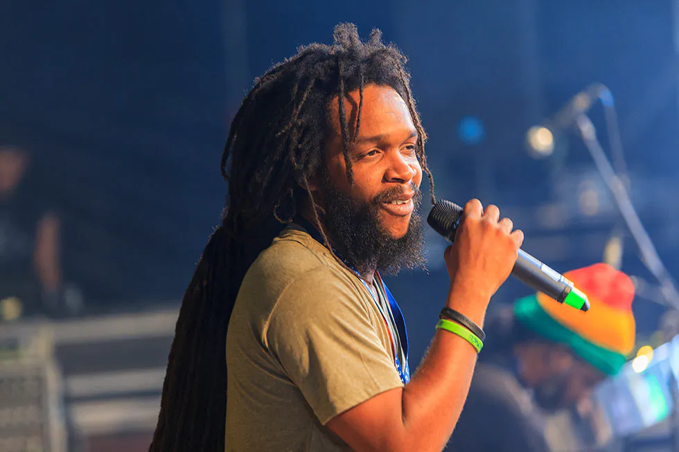 Sänger mit langen Dreadlocks und Bart hält ein Mikrofon auf einer blau beleuchteten Konzertbühne.