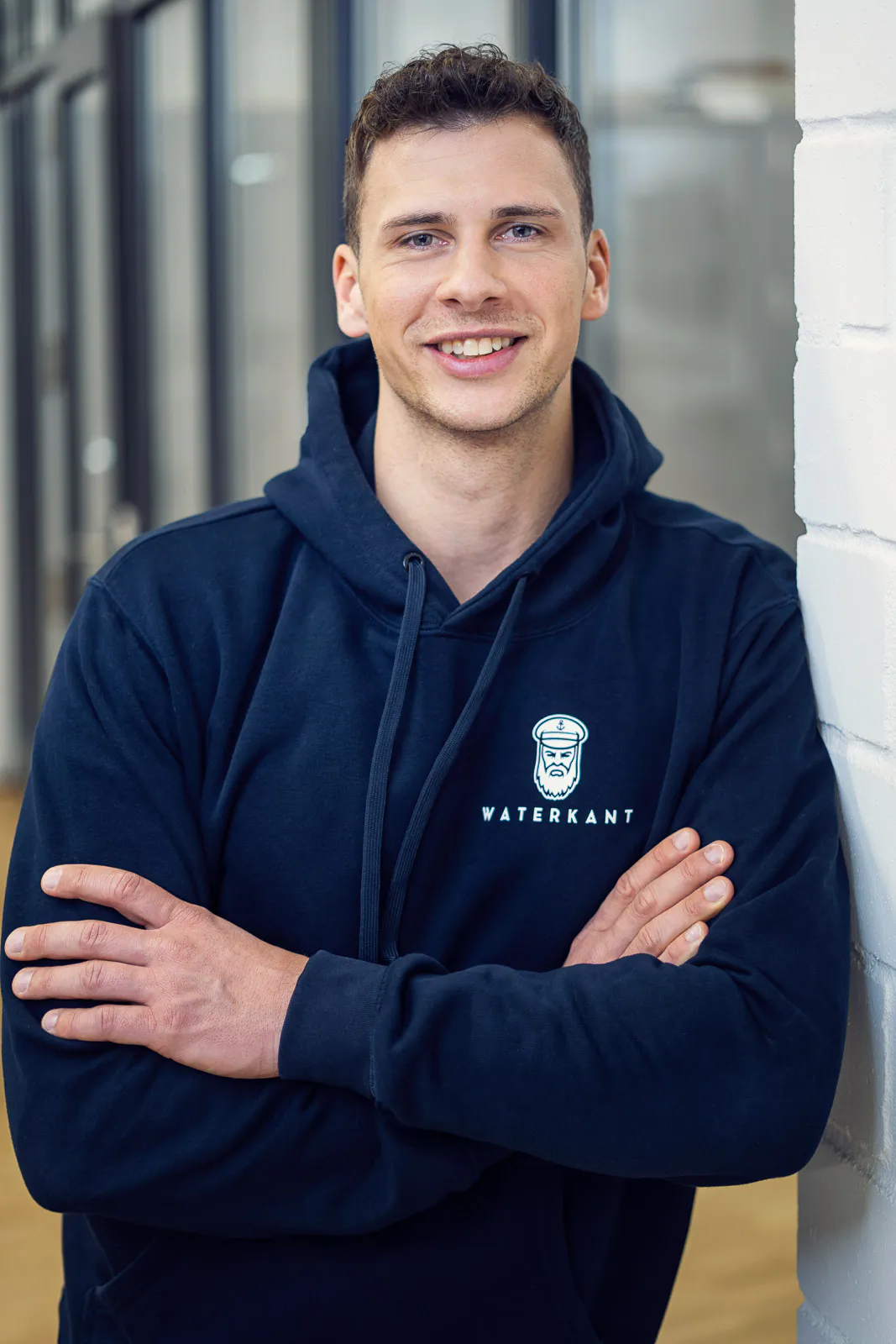 Lächelnder Mann im dunkelblauen Waterkant-Hoodie mit verschränkten Armen vor Fenstern und weißer Backsteinwand.