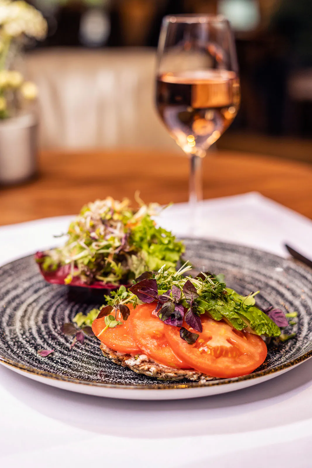 Tomatenscheiben mit Kräutern und Blattsalat auf dunklem Teller, dahinter ein Weinglas im warm beleuchteten Restaurant.